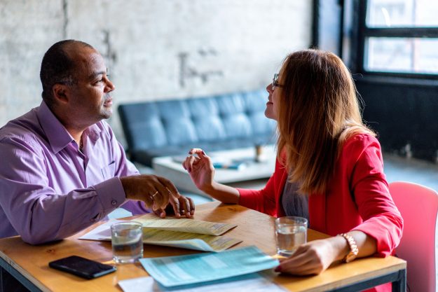 Two business professionals in a discussion, sitting at a table with documents and water glasses, representing collaboration and decision-making during M&A integration.