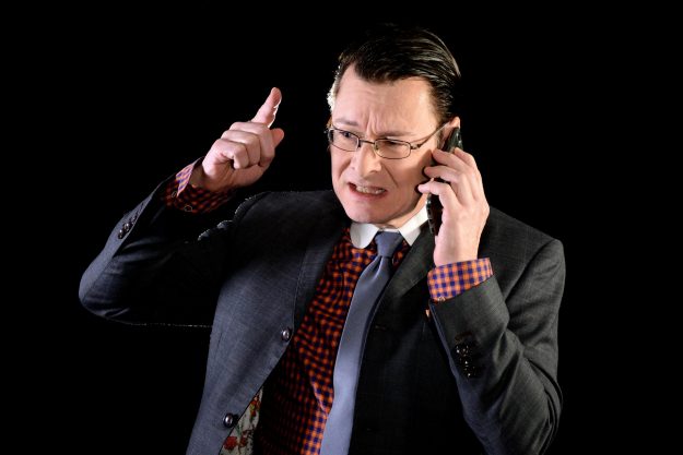 Frustrated businessman in a suit talking on the phone, raising his finger in anger. Represents workplace stress, burnout, and the dangers of overwork in high-pressure environments.