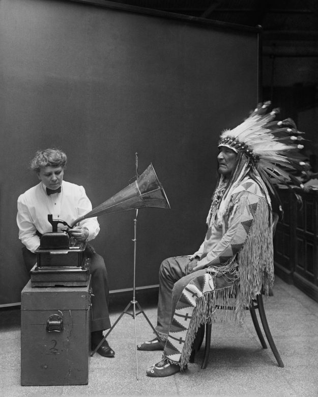 A historical black-and-white photograph of an Indigenous leader wearing a feathered headdress, seated while a woman operates an early phonograph recording device. The image symbolizes tradition meeting modernization.