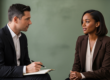 An interviewer in a dark suit takes notes while a candidate in a brown blazer responds during a formal job interview. Article: Recruitment: Buy-and-Build's Most Underpriced Value Accelerator.