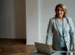 A businesswoman in a light gray blazer stands beside an open laptop in a minimalist modern office with herringbone wood flooring — featured image for "The Missing Pillar of Due Diligence in M&A: Founder Credibility"