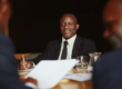 A business leader smiling warmly while engaged in conversation with colleagues at a meeting table — Why the Best Scaling Founders Admit They've Become the Bottleneck